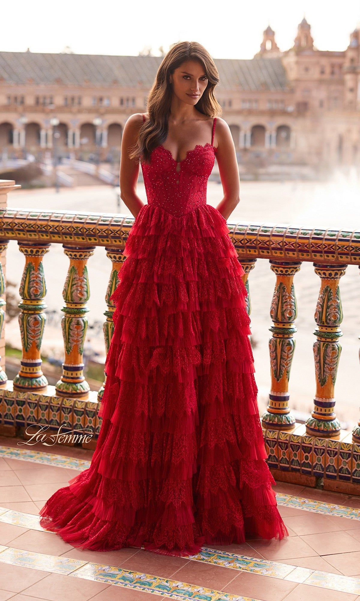 A woman in the La Femme 33525 long prom dress—a tiered red lace gown with a corset bodice—stands on a tiled balcony with ornate railings, overlooking a historic building and water feature.